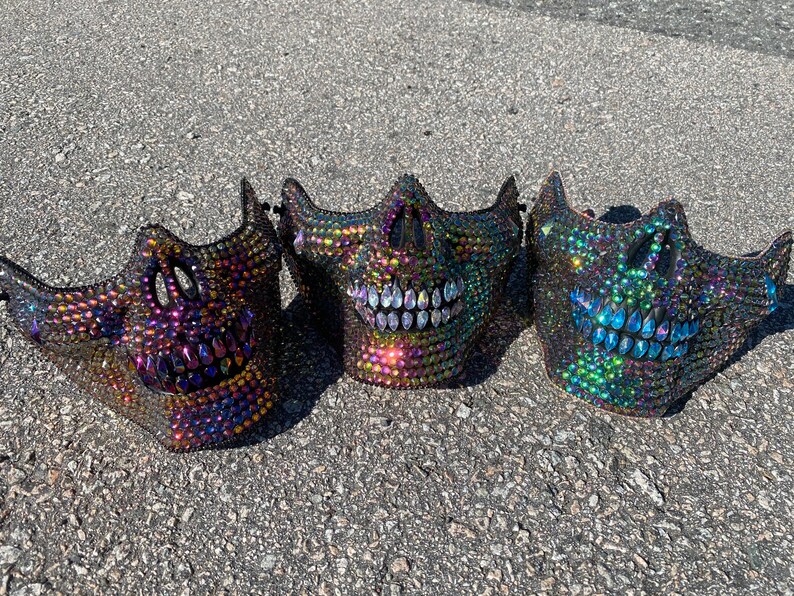 Pu&ograve; includere: Tre maschere di teschio iridescenti con strass. Le maschere sono nere con un arcobaleno di colori negli strass. Le maschere sorridono tutte.