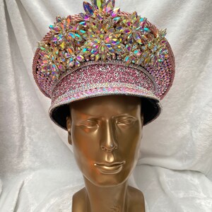 May include: A decorative pink hat with iridescent gemstones and rhinestones. The hat features a wide brim and is adorned with multiple floral-shaped embellishments. The hat is displayed on a gold mannequin head.