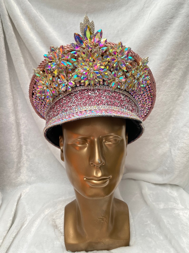 May include: A decorative pink hat with iridescent gemstones and rhinestones. The hat features a wide brim and is adorned with multiple floral-shaped embellishments. The hat is displayed on a gold mannequin head.