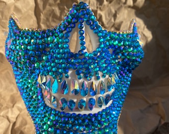 May include: A jeweled face mask in shades of blue and green, with a gold trim. The mask is covered in small, round, and teardrop-shaped rhinestones. The mask has a skeletal design with a row of teeth.
