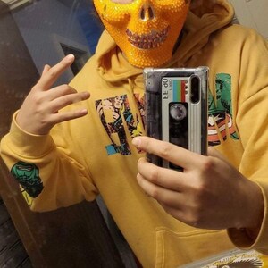 Op de afbeelding: Een persoon draagt een gele hoodie met een print van een stripfiguur en een schedelvormig masker. De persoon houdt een telefoonhoesje vast met een cassettebandontwerp. Het telefoonhoesje is transparant en laat het telefoonscherm zien.