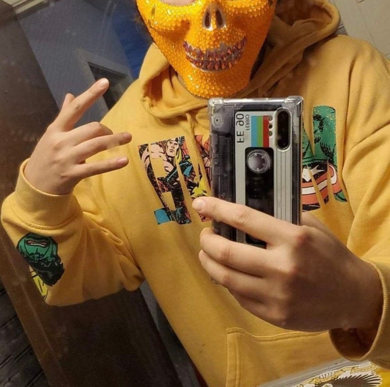 Op de afbeelding: Een persoon draagt een gele hoodie met een print van een stripfiguur en een schedelvormig masker. De persoon houdt een telefoonhoesje vast met een cassettebandontwerp. Het telefoonhoesje is transparant en laat het telefoonscherm zien.