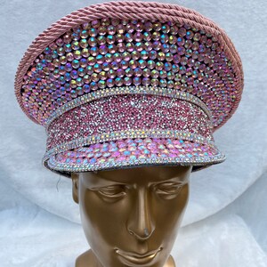 May include: A pink, bedazzled hat with iridescent rhinestones. The hat features a circular crown, a flat brim, and a decorative rope trim. The hat is displayed on a gold mannequin head.