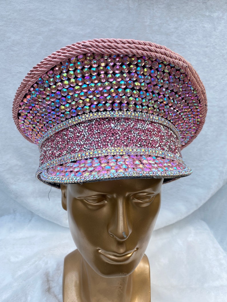 May include: A pink, bedazzled hat with iridescent rhinestones. The hat features a circular crown, a flat brim, and a decorative rope trim. The hat is displayed on a gold mannequin head.