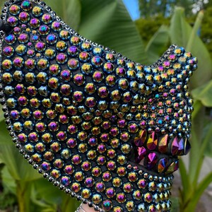 May include: A black face mask densely covered with iridescent, round, and faceted rhinestones. The mask has a silver beaded trim and features a stylized jawline with pointed, colorful crystal accents. The mask is held up by a hand.