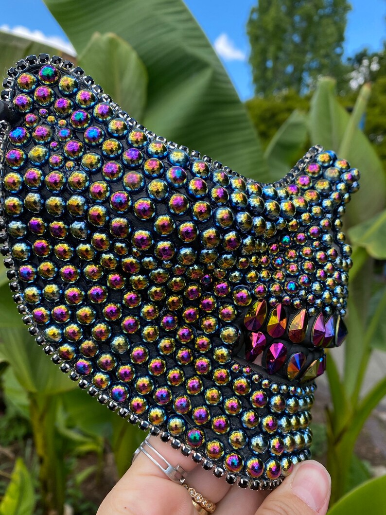 May include: A black face mask densely covered with iridescent, round, and faceted rhinestones. The mask has a silver beaded trim and features a stylized jawline with pointed, colorful crystal accents. The mask is held up by a hand.