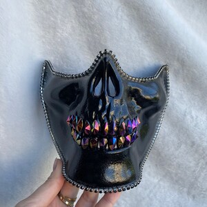 May include: Black skull mask with iridescent rhinestones for the teeth. The mask is made of a shiny, black material and has a beaded trim.