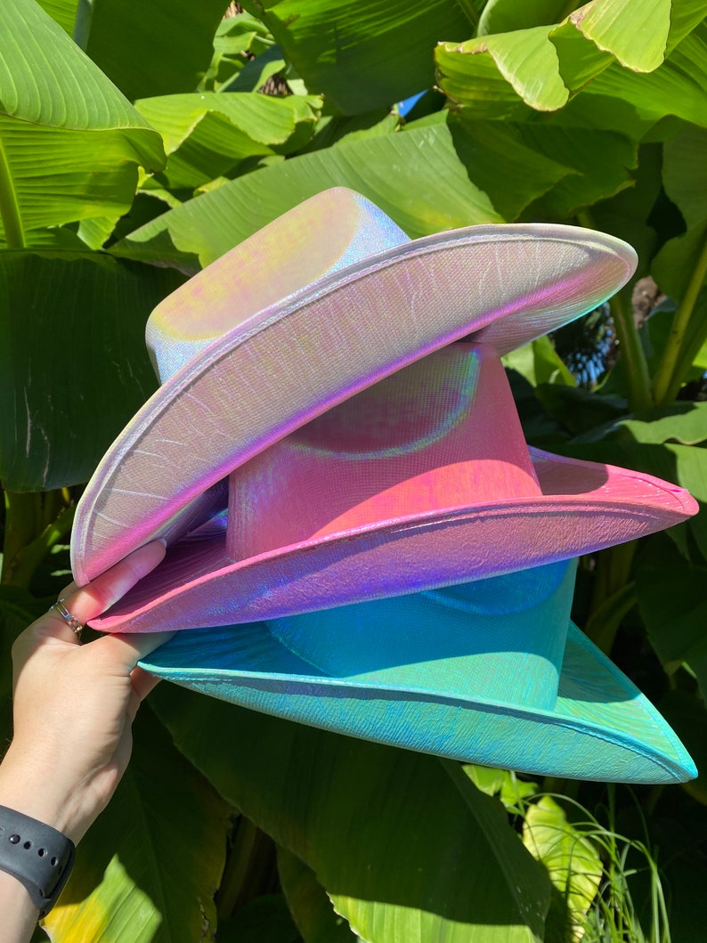 May include: Three iridescent cowboy hats stacked on top of each other. The top hat is a light purple, the middle hat is pink, and the bottom hat is a light blue.