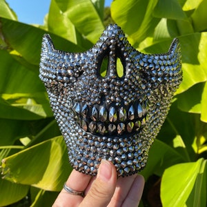 Puede incluir: Una máscara de calavera negra cubierta de pedrería plateada. La máscara tiene un diseño puntiagudo y espinoso.