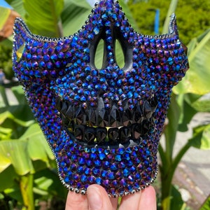 May include: A black skull mask covered in iridescent blue and purple rhinestones. The mask has a detailed design with a black mouth and teeth.