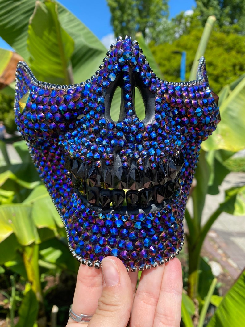 May include: A black skull mask covered in iridescent blue and purple rhinestones. The mask has a detailed design with a black mouth and teeth.