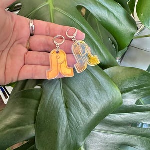 May include: A pair of orange and yellow acrylic earrings with a unique, abstract design. The earrings are held up against a backdrop of large, green leaves. The earrings have silver-colored ear wires.