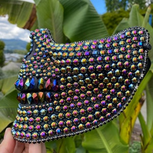 May include: A black mask covered in iridescent rhinestones. The mask has a jagged, tooth-like design on the lower part and is outlined with silver beads. The rhinestones reflect a rainbow of colors.