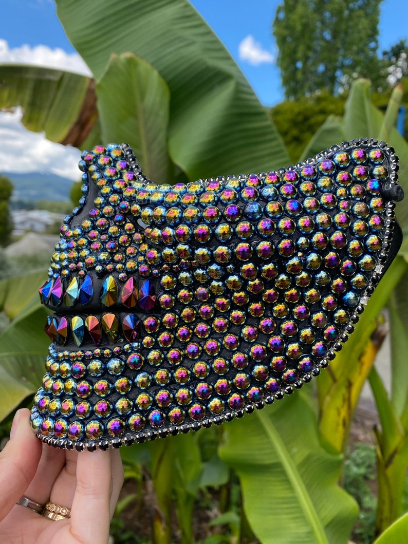 May include: A black mask covered in iridescent rhinestones. The mask has a jagged, tooth-like design on the lower part and is outlined with silver beads. The rhinestones reflect a rainbow of colors.