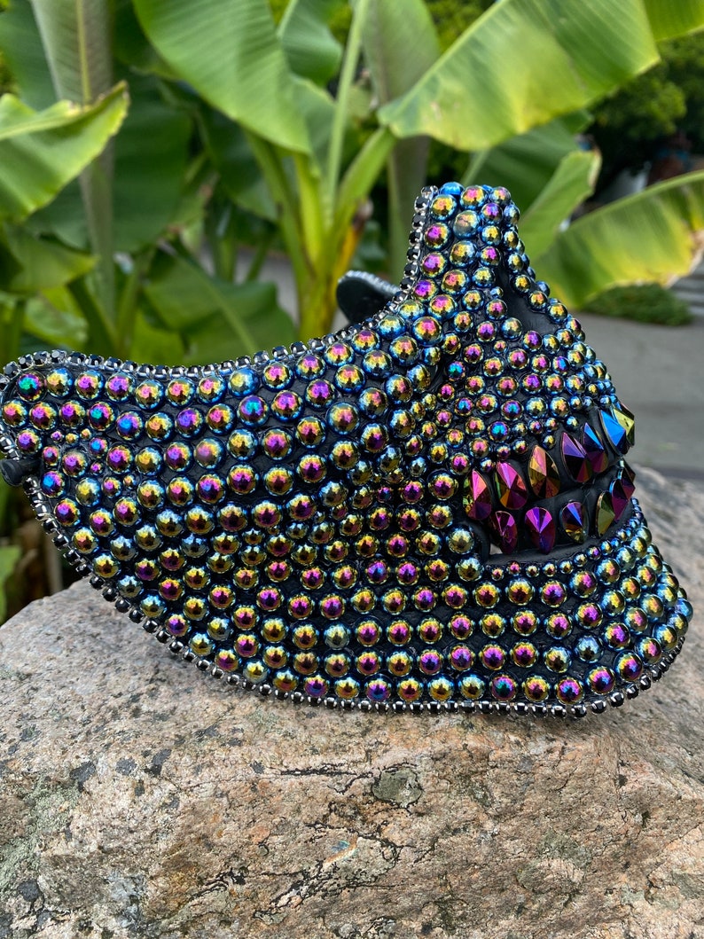 May include: A black mask covered in iridescent, round, and pointed rhinestones. The mask has a curved shape with a pointed top and a jagged, open mouth design. The edges are lined with small silver beads.