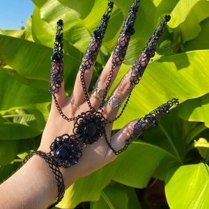 May include: Black metal finger rings with intricate designs and black gemstones. The rings are connected by chains and form a bracelet on the wrist.