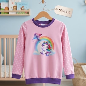 Könnte beinhalten: Ein rosa Langarm-Sweatshirt mit lila Kragen und Bündchen. Die Ärmel haben ein weißes Sternmuster. Auf der Vorderseite ist ein Cartoon-Einhorn mit einem Drachen vor einem Regenbogen abgebildet. Ein Etikett zeigt "Size 116".