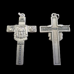 May include: A silver cross pendant with religious iconography. The front shows a detailed image of Jesus and the inscription "SIT NOMEN DOMINI BENEDICTUM". The reverse side has the text "VADE RETRO SATANA!"