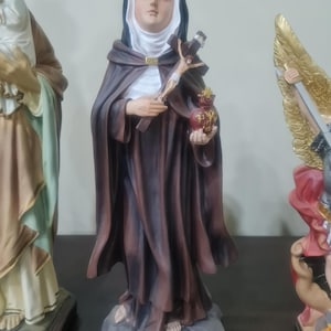 Könnte beinhalten: Eine Statue der heiligen Veronika Giuliani, die einen dunkelbraunen Umhang und einen weißen Schleier trägt und ein Kruzifix und einen herzförmigen Gegenstand hält. Die Statue steht auf einem grauen Sockel mit Text. Andere Statuen sind teilweise sichtbar.