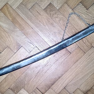 May include: A silver-colored sword with a chain attached to the handle. The sword is lying on a wooden floor.