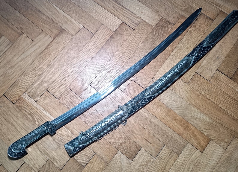 May include: A silver and black sword with an ornate handle and scabbard. The sword has a curved blade and intricate engravings on the scabbard.