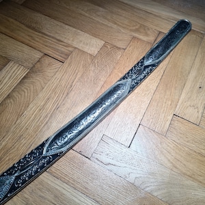 May include: A silver sword with an intricate floral design on the blade. The sword is lying on a wooden floor.