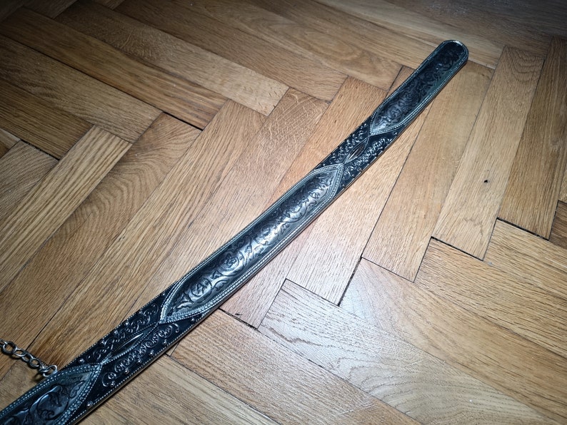 May include: A silver sword with an intricate floral design on the blade. The sword is lying on a wooden floor.