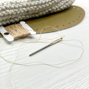 May include: A close-up of craft supplies including a silver needle, beige thread, a spool of thread, a light gray knitted item, and a tan leather base with pre-punched holes. The items are arranged on a white surface, suggesting a crafting project.