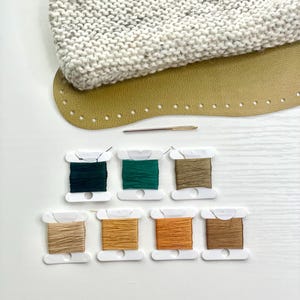 Puede incluir: Un kit de manualidades con un artículo de punto gris, una base de cuero marrón claro con agujeros para coser y carretes de hilo de varios colores: verde oscuro, verde, marrón claro, naranja y marrón. También se incluye una aguja.