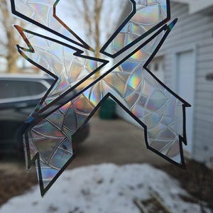 May include: A holographic sticker with a black outline of the letter X. The sticker is on a window with a view of a snowy yard and a house in the background.