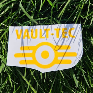 May include: A white sticker with the text "VAULT-TEC" in yellow letters and a yellow Vault-Tec logo with a white circle in the center.