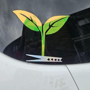 May include: A green and yellow iridescent vinyl sticker of a plant with two leaves and a stem. The plant is growing out of a pair of silver scissors.