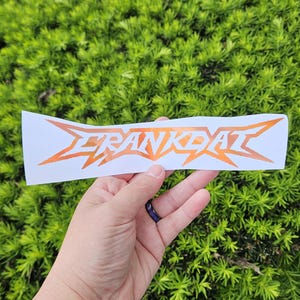 May include: A white sticker with the word "FRANKDAT" in orange, stylized lettering. The sticker is held against a backdrop of green foliage. The text has a jagged, angular design.