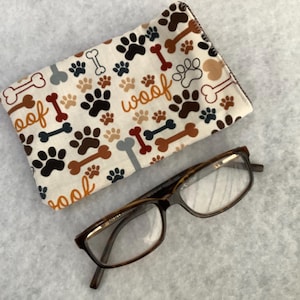 May include: A white fabric eyeglass case with a paw print and bone pattern. The case has the word "woof" printed on it in orange. A pair of brown-framed spectacles are lying next to the case.