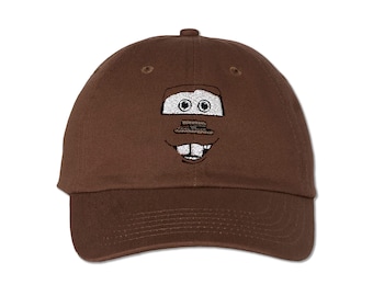 May include: A brown baseball cap featuring an embroidered cartoon face. The design includes white eyes, a rectangular mouth, and a smiling expression. The cap has a curved brim and a fabric strap closure.