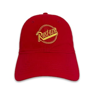 May include: A red baseball cap with a curved brim. The cap features the embroidered text "Rusteze" in yellow within a yellow circle. The hat is made of a soft fabric and has a button on top.