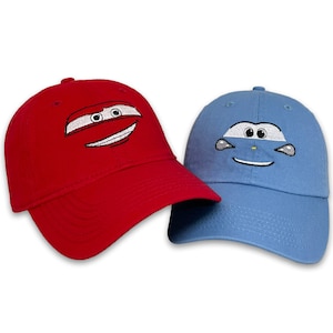 May include: Two baseball caps, one red and one light blue, each embroidered with a cartoon car face design. The red cap has a smiling face, while the blue cap has a neutral expression. Both caps have a curved brim and a top button.