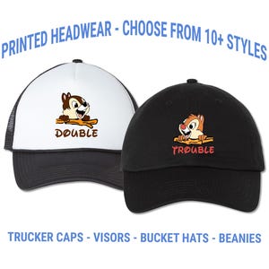 May include: Two baseball caps, one black and one white, with cartoon chipmunk graphics. The white cap has a black mesh back and the word "DOUBLE". The black cap has the word "TROUBLE". Text at the top says "PRINTED HEADWEAR - CHOOSE FROM 10+ STYLES".