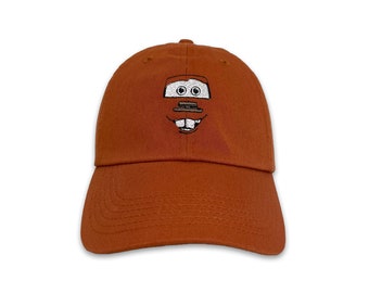 May include: A rust-colored baseball cap featuring an embroidered cartoon face. The design includes two eyes, a rectangular mouth, and a smiling expression. The cap has a curved brim and a six-panel construction.