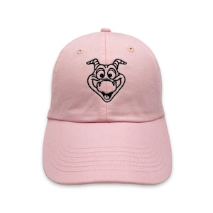 May include: A pale pink baseball cap with a black embroidered cartoon face. The cap has a curved peak and an adjustable strap at the back. The design is centered on the front of the cap.