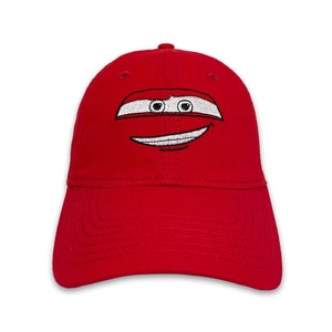 May include: A bright red baseball cap featuring an embroidered cartoon face with white and black details. The design includes a white stripe with black eyes and a smiling mouth. The cap has a curved brim and a rounded crown.