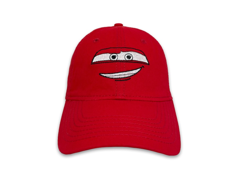 May include: A bright red baseball cap featuring an embroidered cartoon face with white and black details. The design includes a white stripe with black eyes and a smiling mouth. The cap has a curved brim and a rounded crown.