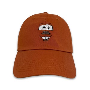 May include: A rust-colored baseball cap featuring an embroidered cartoon face. The design includes two eyes, a rectangular shape, and a smiling mouth. The cap has a curved brim and a fabric strap closure.