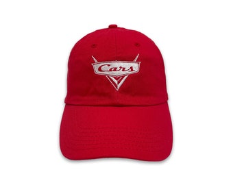 May include: A red baseball cap featuring the word "Cars" in white embroidery. The logo is a stylized emblem with a lightning bolt design. The cap has a curved brim and an adjustable strap.