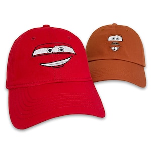 May include: Two baseball caps, one red and one brown, with embroidered cartoon car faces. The red cap features a smiling face with white and black details. The brown cap has a different car face design. Both caps have a curved bill and a top button.