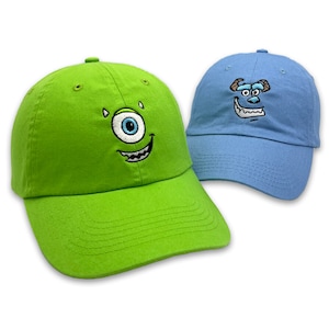 May include: Two baseball caps, one lime green and one light blue, each embroidered with a cartoon monster face. The green cap features a one-eyed monster, while the blue cap has a two-eyed monster design. Both caps have a curved brim and a top button.