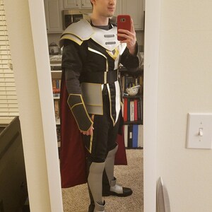 May include: A person wearing a black and silver costume with a red cape. The costume features a silver breastplate with a gold trim and a black belt. The person is holding a red phone in their right hand.