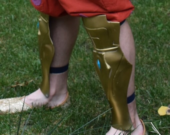 May include: Gold-colored leg armor with a blue gem detail. The armor is worn over a pair of brown sandals.