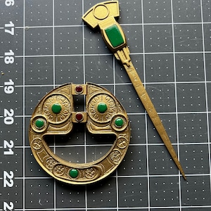 May include: A gold-colored Celtic knot brooch with green and red accents, and a matching gold-colored pin with a green accent.