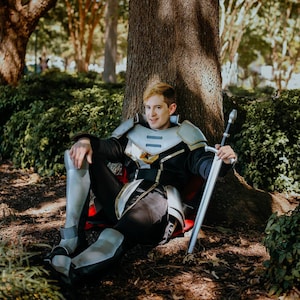 May include: A person wearing silver armor and a black tunic sits against a tree with a sword in their hand. The person is looking at the camera and smiling.